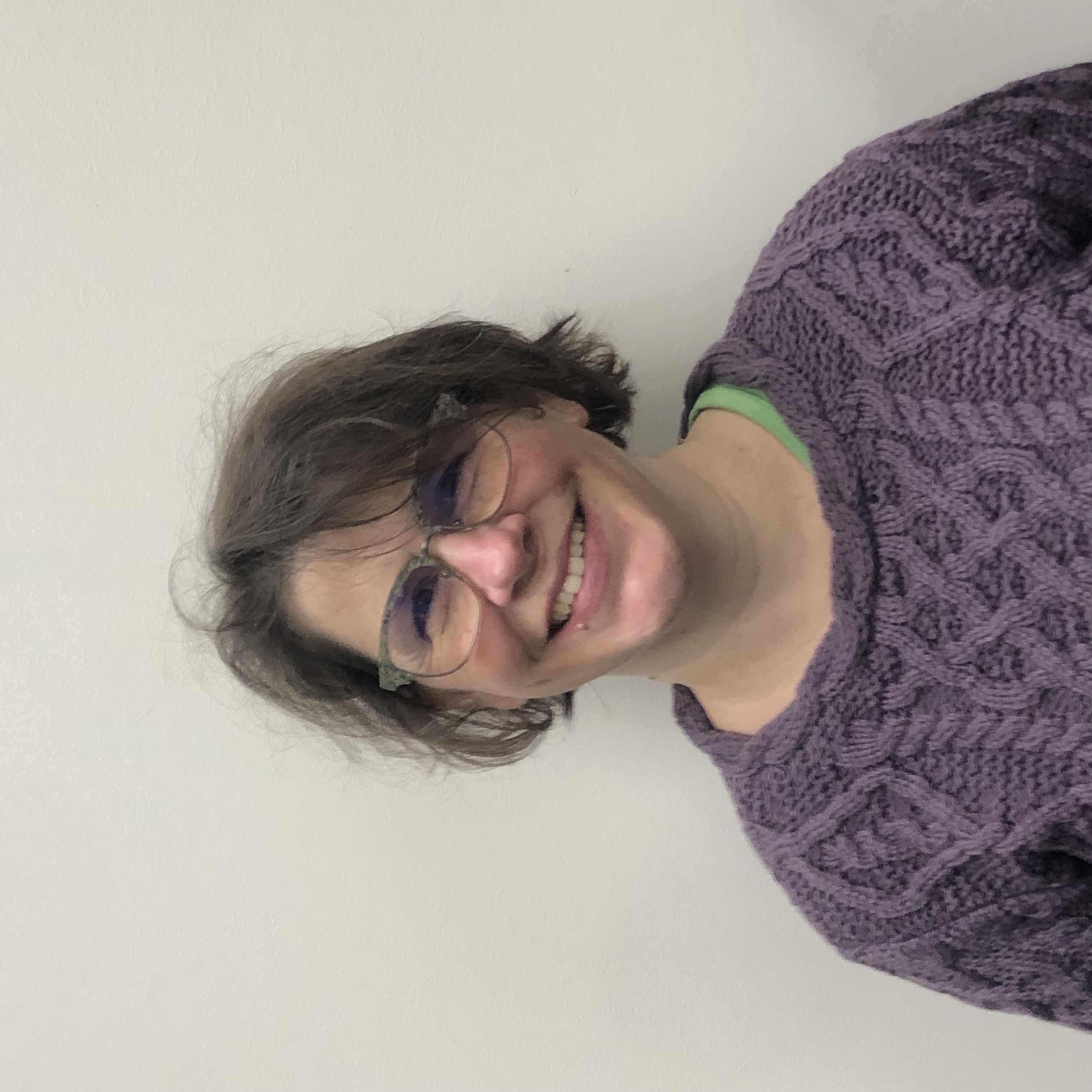 Jacy Pietsch - a woman with short-hair swooping over her left eye smiles at the camera. She wears glasses and a purple mock turtleneck sweater. 