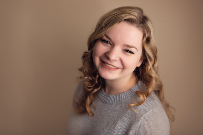 A woman with light brown loosely curled hair smiles up at the camera. She wears a light gray crewneck sweater. The background is light brown.