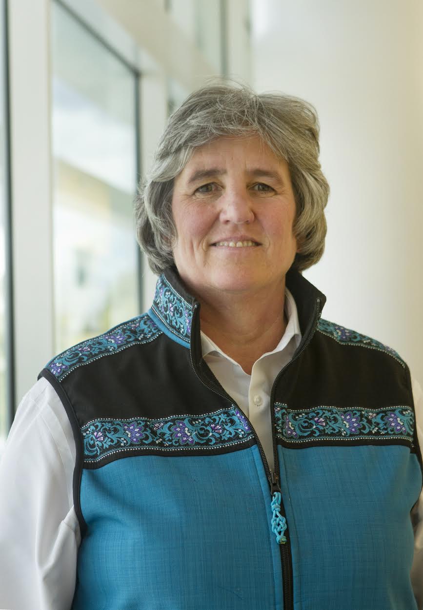 A woman with short hair (UA President Pat Pitney) wearing a teal vest stands in a well-lit hallway smiling at the camera.