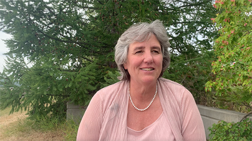 Pat Pitney, a gray-haired woman with short hair, sits outside amidst greenery. She wears a pink sweater and pearl necklace as she smiles at something in the distance.