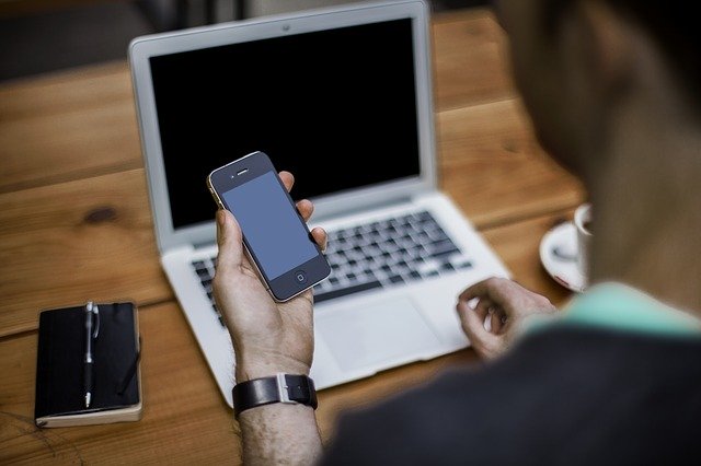 A person holds a cell phone in front of a table with a laptop and notebook on it