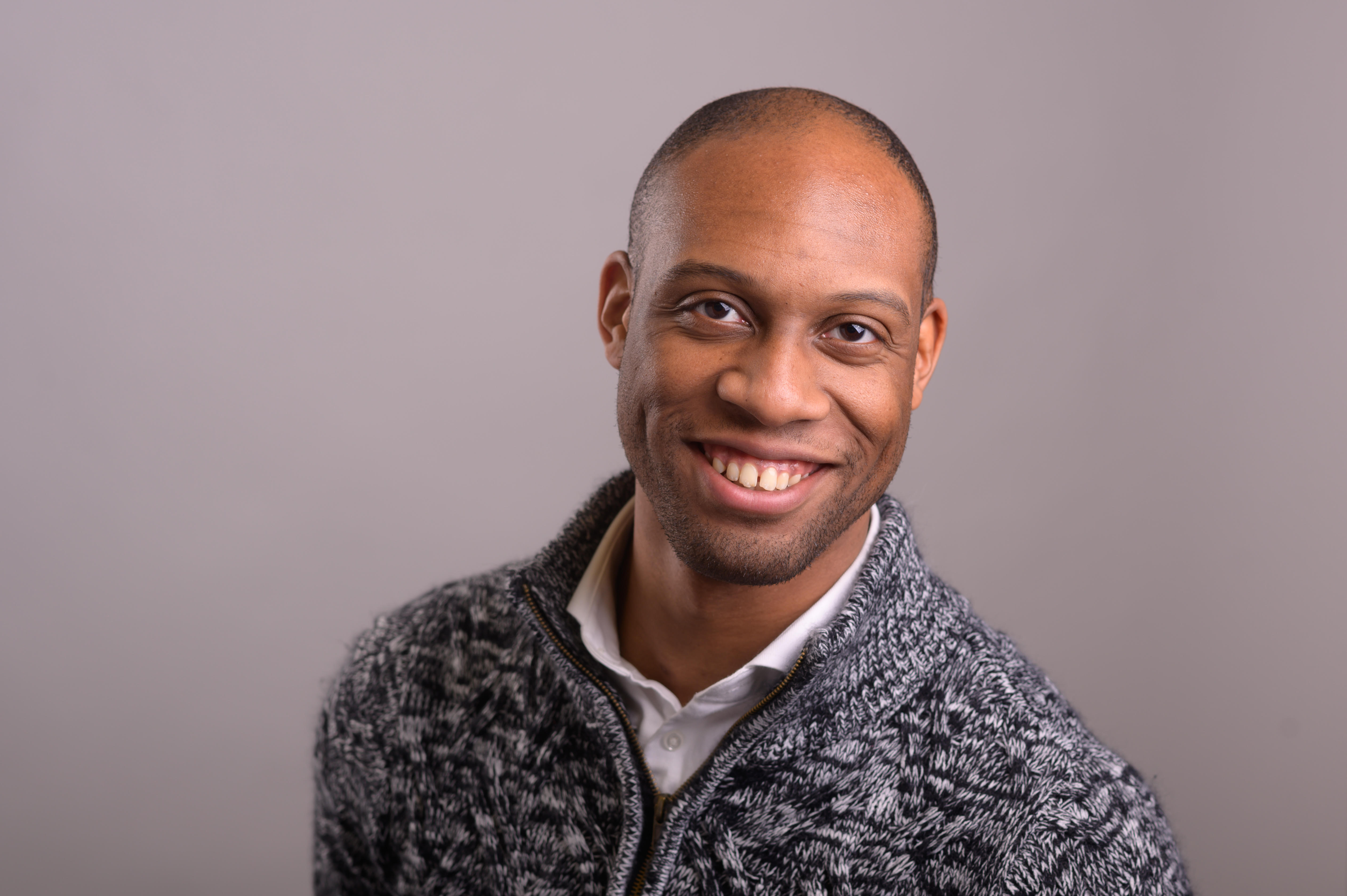 Jonathon Taylor, a person with buzzed hair and a sweater over a white button up shirt, smiles at the camera