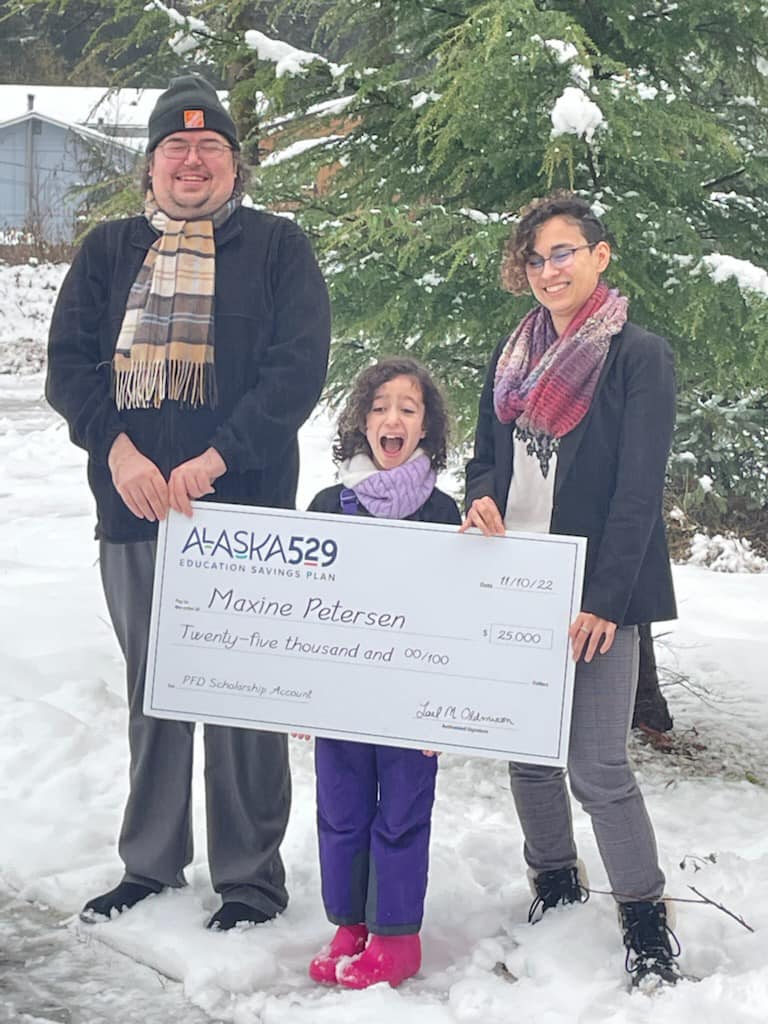 Maxine Peterson, a small girl, stands in the snow with two adults. All three people are smiling, Maxine's mouth is wide open-happy. She holds a giant novelty check for $25,000 from Alaska 529.