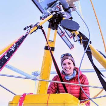 Emily Fedders smiling in a red jacket among yellow research equipment outdoors.