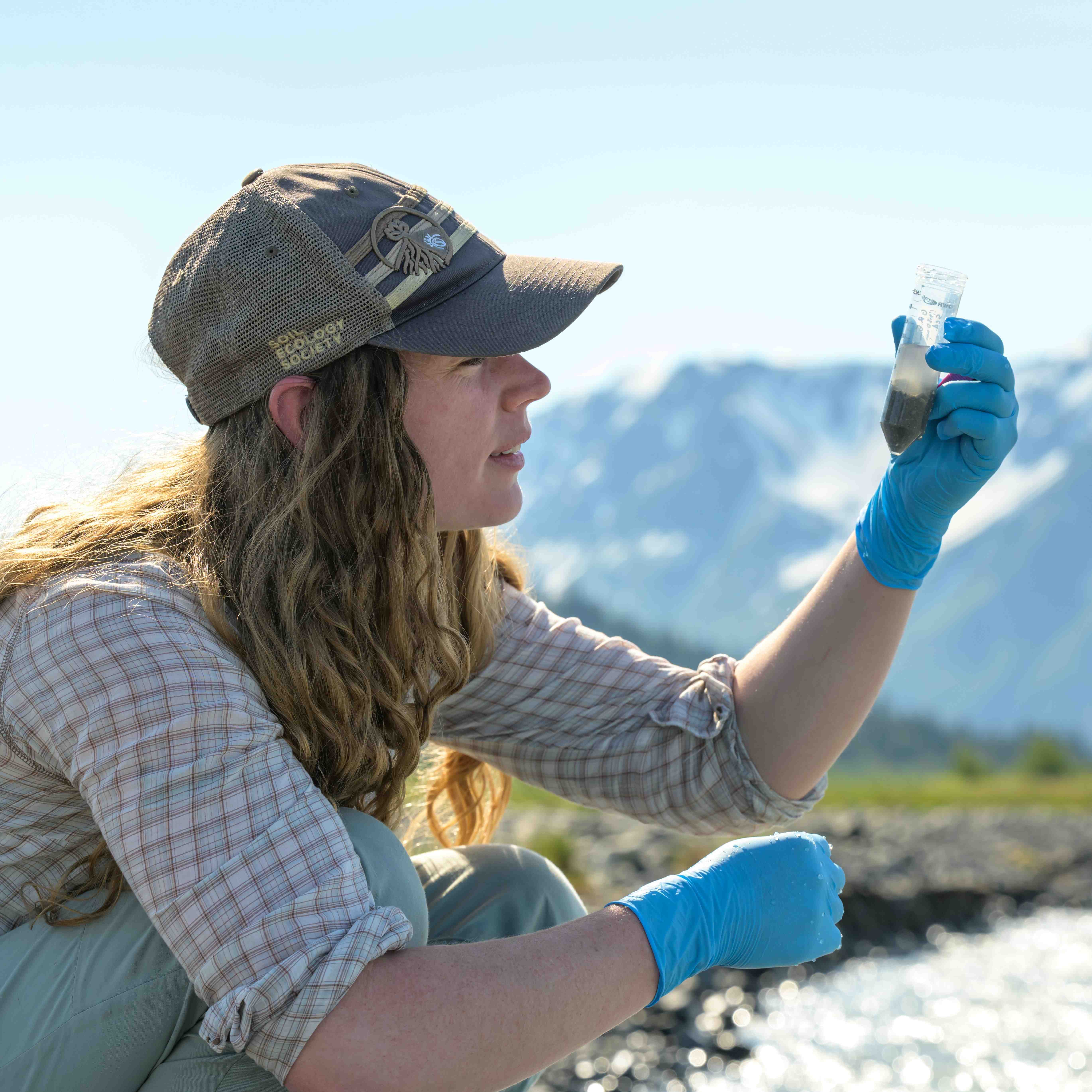 Memphis Hill wearing gloves and a cap, examining a sample vial by a river with mountains in the background.