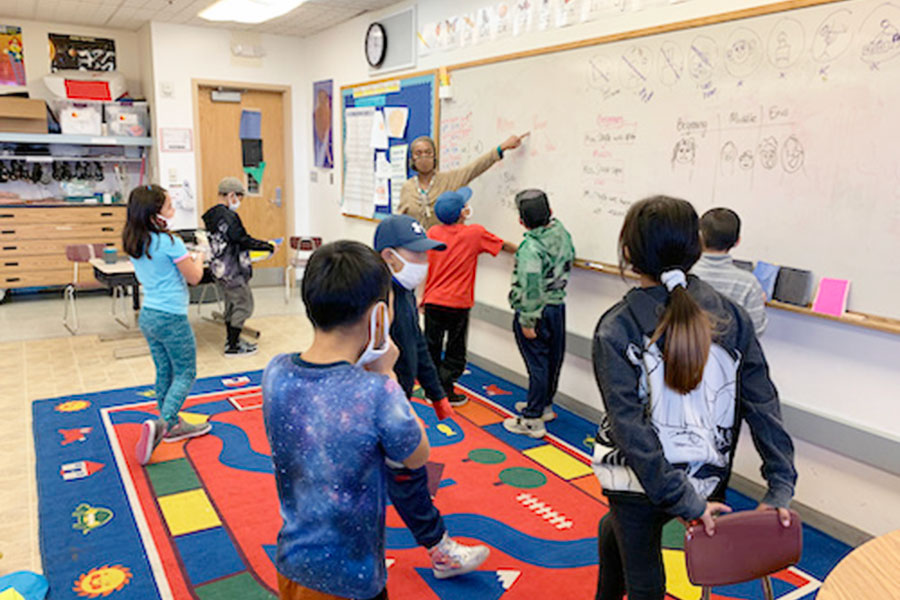 A teacher points to the whiteboard in an elementary classroom as children look on