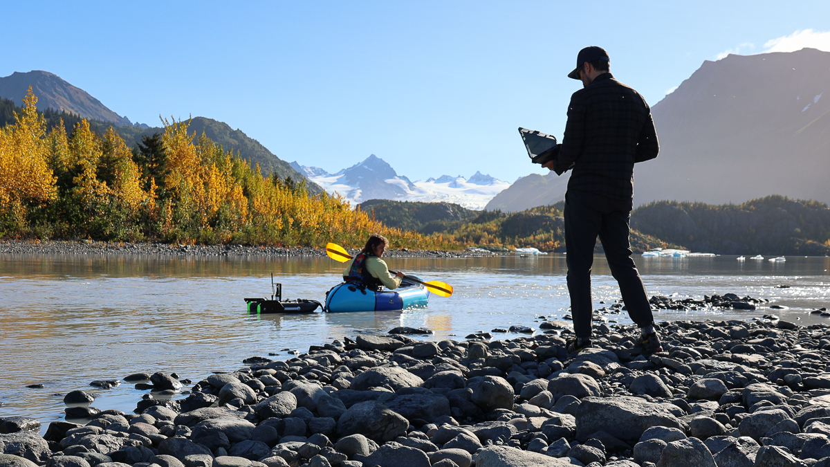 Jordan stands tall and silhouetted against the scene of a glacial stream. Aeon paddles the packraft in the stream. It is a gorgeous sunny fall day.
