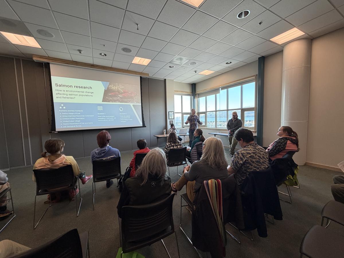 A speaker giving a presentation in a sunlit room full of people of mixed ages.