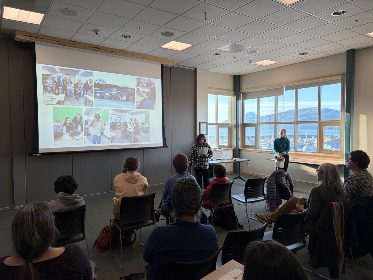 A speaker giving a presentation in a sunlit room full of people of mixed ages.