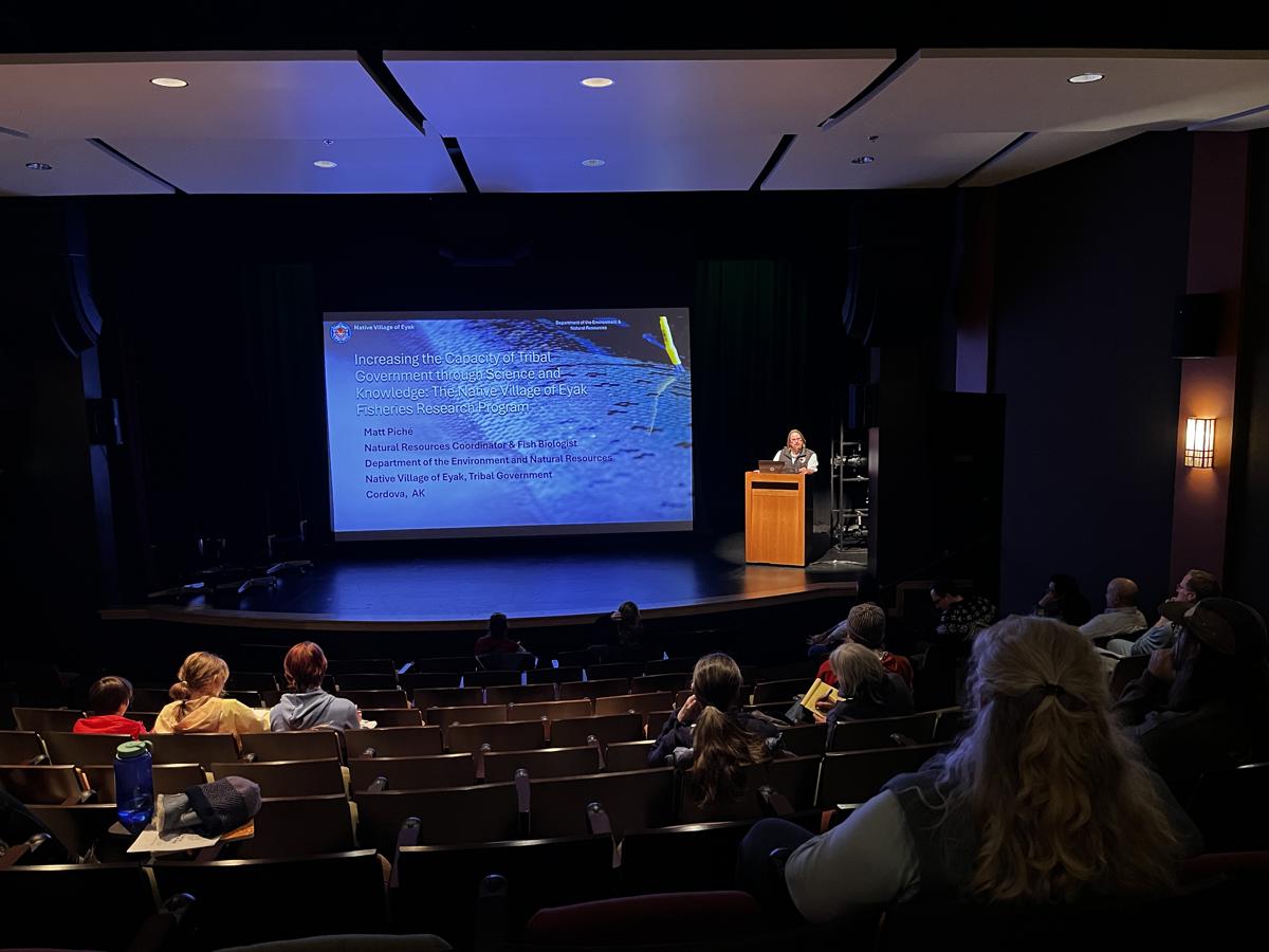 A speaker giving a presentation in a sunlit room full of people of mixed ages.