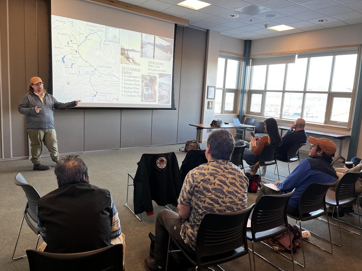 A speaker giving a presentation in a sunlit room full of people of mixed ages.