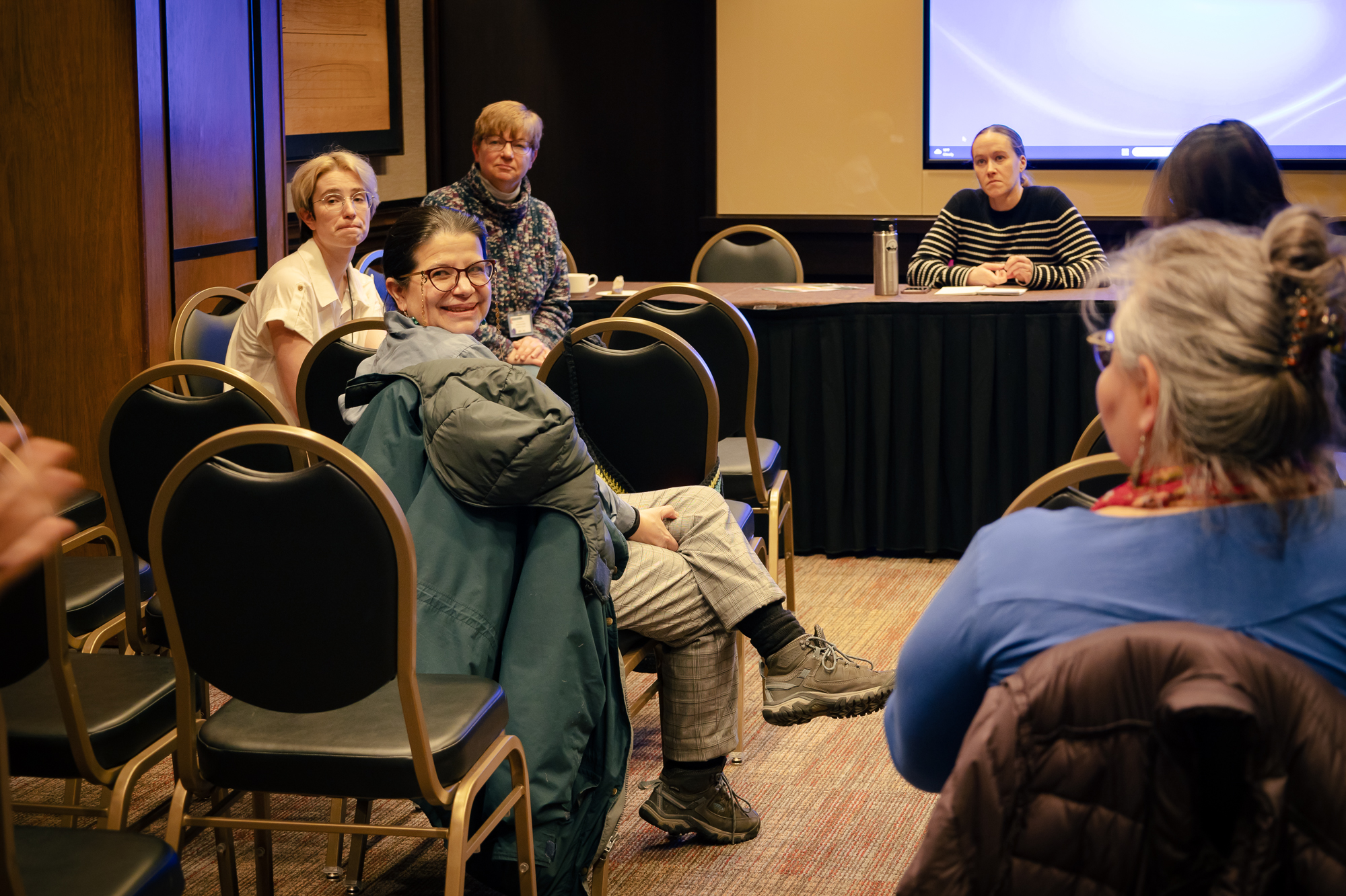 Women in a panel presentation audience a conference room turned around listening and smiling.