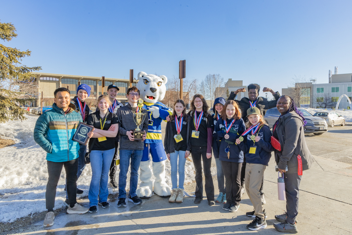 A middle school team group photo with the UAF Nanook on a clear blue sunny winter day in late March.
