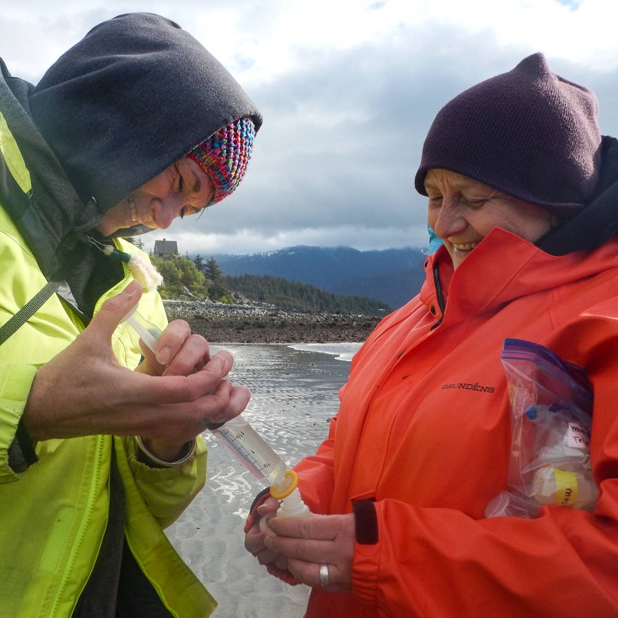 Two women smiling and helping each other collect a water sample on a beach in Southeast Alaska.