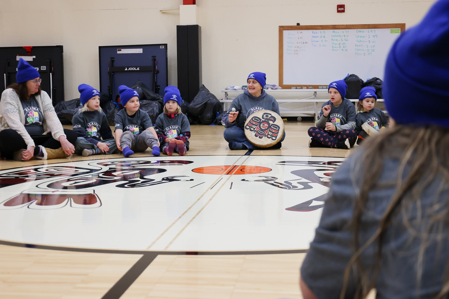 “We listen with our ears,” said Justina Starzynski-Hotch as she tugged on her earlobe. She said the phrase in Tlingit first, then in English as she sat in a circle of 33 children in the center of the gymnasium of the Klukwan School. Their cross-legged bodies circled around the school’s logo: a formline representation of Eagle and Raven, the two Tlingit moieties, leaping after a basketball.