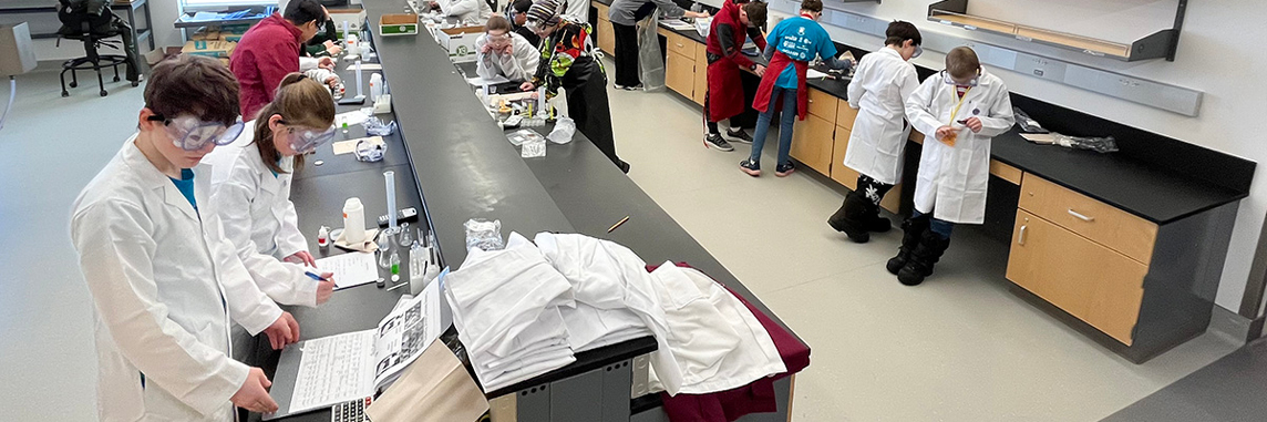 Middle school students wearing white lab coats and safety goggles puzzle over chemistry equipment and notes in a lab.