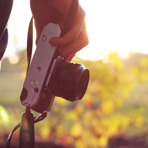 A hand holding a camera with fall foliage blurred in the background.