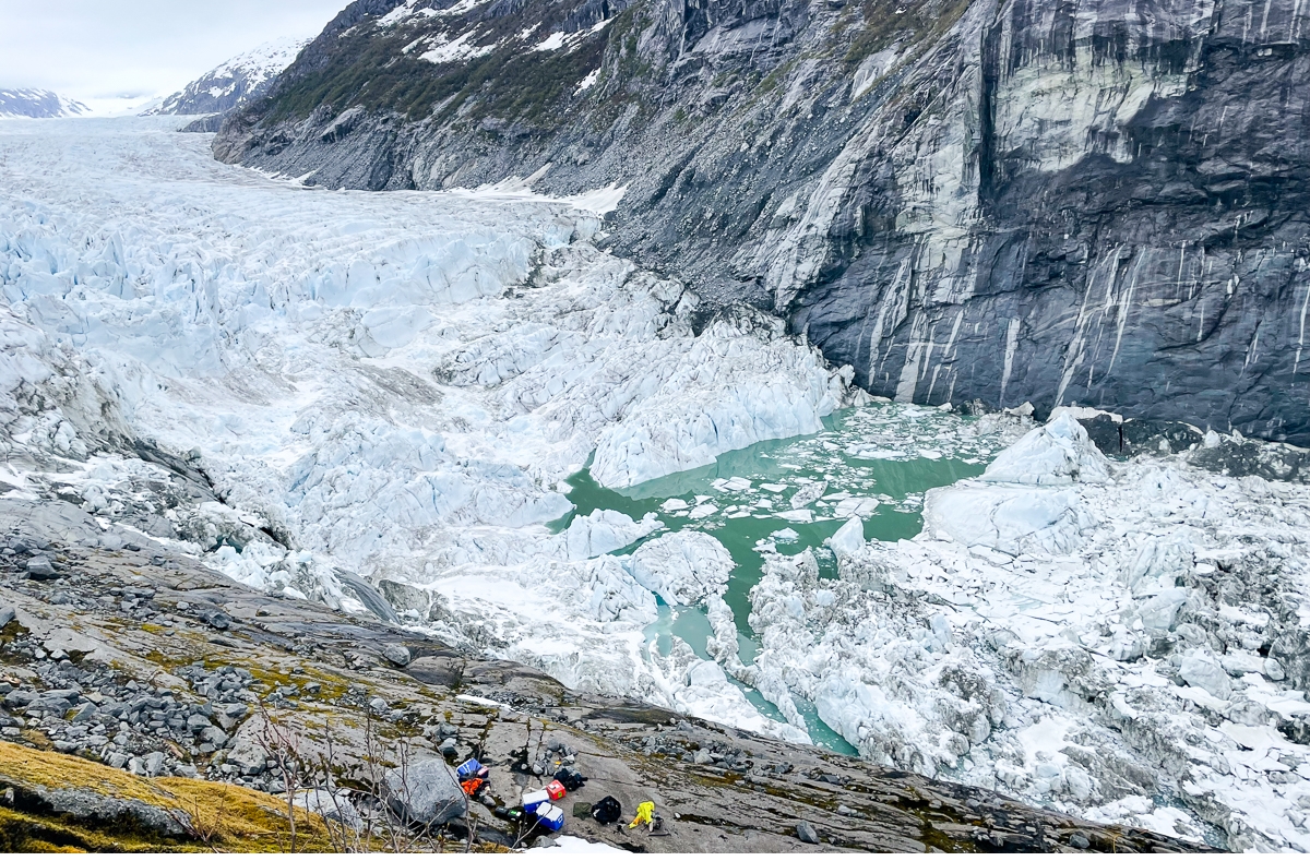 A majestic, somewhat dreary scene of a glacier taken from mossy mountain rock beside it. The ice is breaking a apart, and a body of teal water has pooled, waiting to flood. There are field work tools and packs in the foreground.