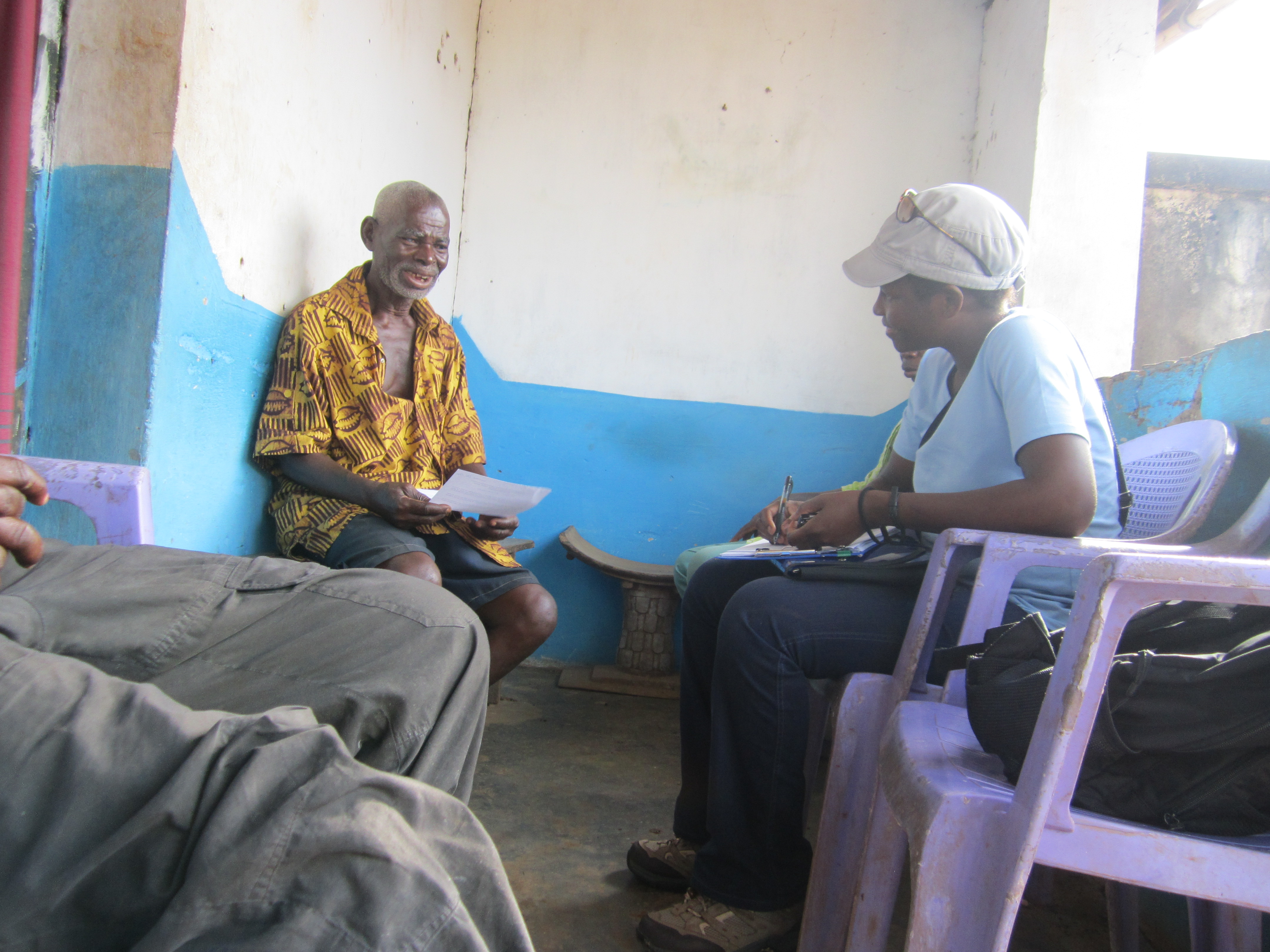 Josie Sam sits in one of three purple chairs. The paint on the chairs is peeling. Josie holds a clip board and a pen, slightly smiling, leaning in and listening to a man seated opposite of her. Sam is wearing a brimmed hat with sunglasses on top of the hat, a white t-shirt, jeans, and hiking boots. The man sits against the wall. He is holding a paper. He may be smiling a little or he is just talking. He wears shorts and a bright yellow short-sleeved, buttoned, collared shirt with a brown pattern. The shirt is unbuttoned at the top, revealing some of his chest, suggesting a hot environment. Lines in his face suggest he is maybe middle-aged, but not elderly. A third person sits in another of the purple chairs, but all we can see is their knees and fingers. The room they are in is small. The thick concrete walls are painted white on top and bright blue on the bottom up to window height. The paint is a bit textured with age. A large window, which is an open section of wall -- no glass or screen, allows light in, washing out the photo somewhat.