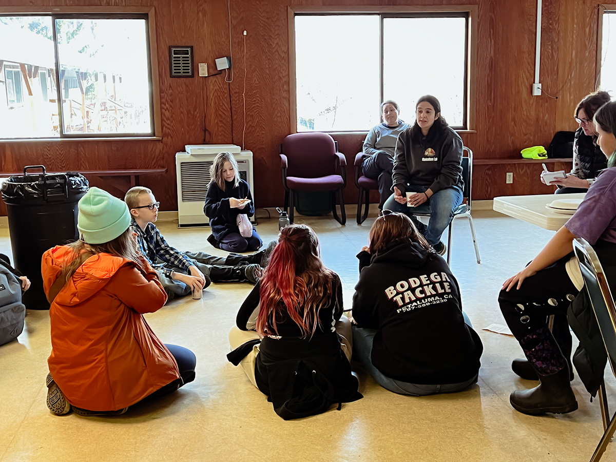 Kids sitting on the floor in a circle with an adult sitting in a chair, backlit by bright windows.