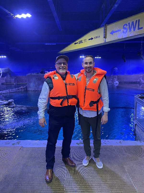 Two men pose for a photo wearing life jackets in front of a pool with large boulders around it.