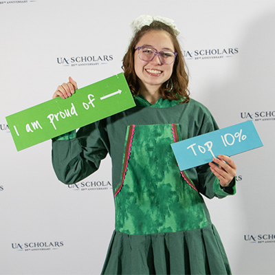 A UA Scholar photo booth portrait holding signs saying top 10% and "I'm proud of" with an arrow.