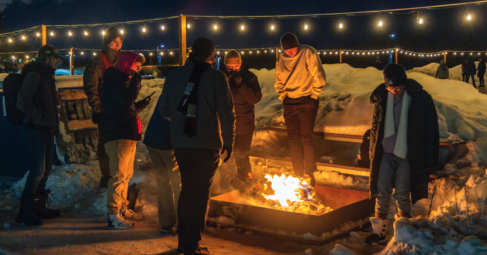 Students at UAF stand around a campfire with the lighted ice skating arena behind them.