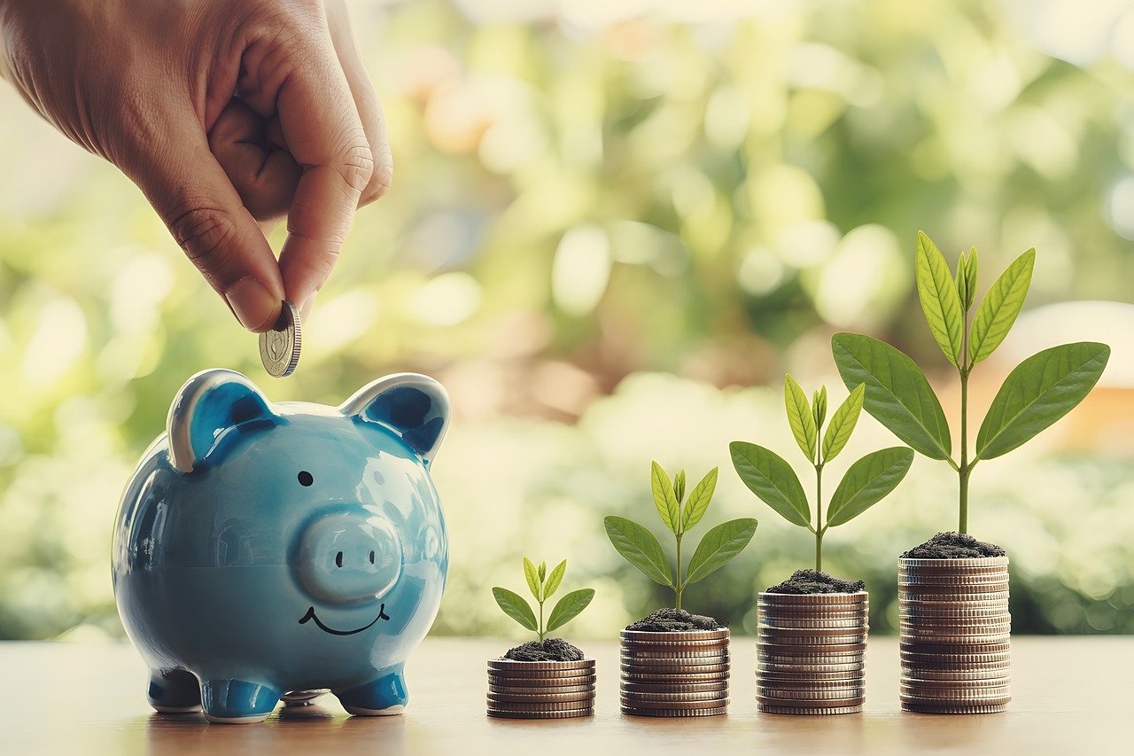 Picture of a hand putting a coin into a small blue piggy bank with a stack of coins next to the piggy bank indicating financial growth