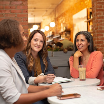 Three women sitting around two small round tables, two women are paying attention to the third.