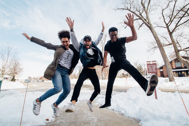 Three people jumping in air with arms spread out and happy expressions on a pathway with snow on the ground.