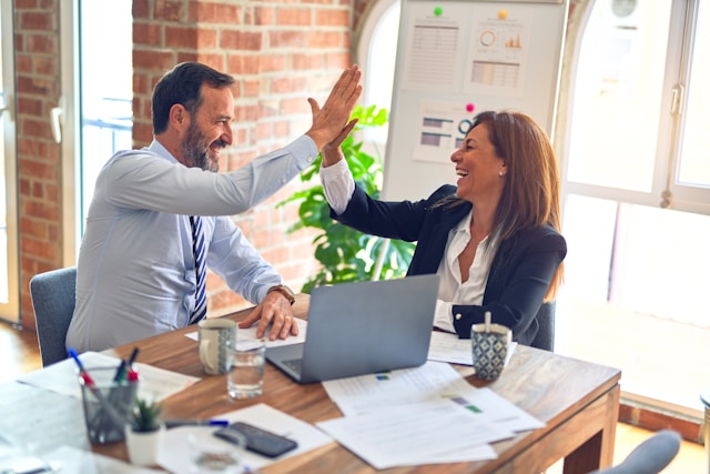 A mand and a woman in an office setting high five over a laptop.