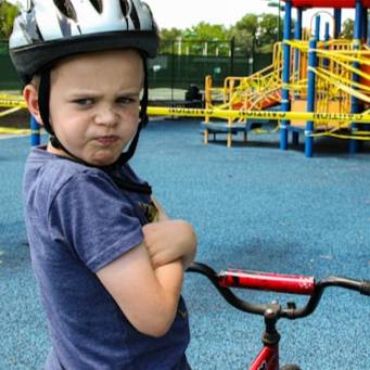 A young boy wearing a bike helmet and a blue shirt stands with a pouty expression next to his bicycle in front of a playground.