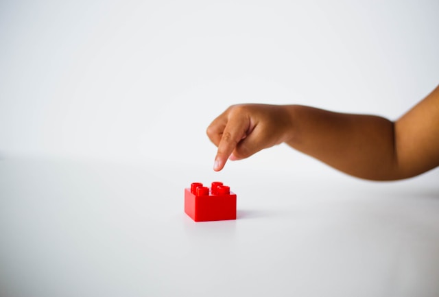 A baby’s arm extending out to the side and pressing a finger on a red building block against a white background