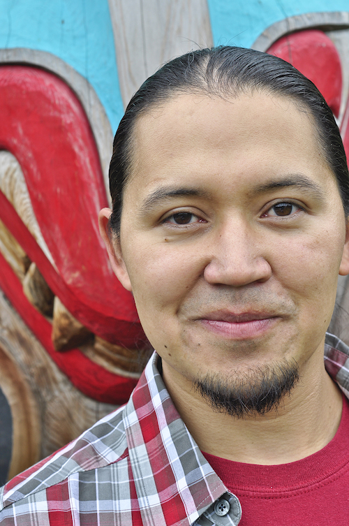A man with a bead smiles in front of a red and blue totem pole