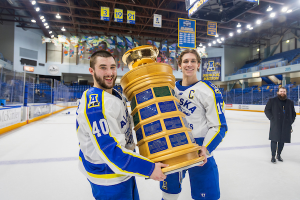Two hockey players stand on the ice rink holding a large trophy