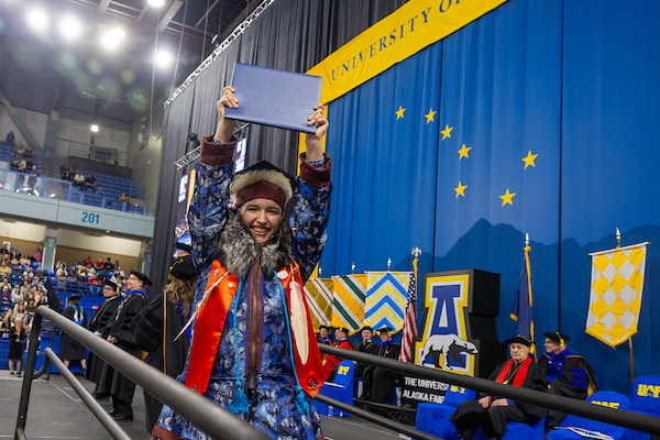 a graduate in Indigenous garb holds up their blue diploma after walking across the stage