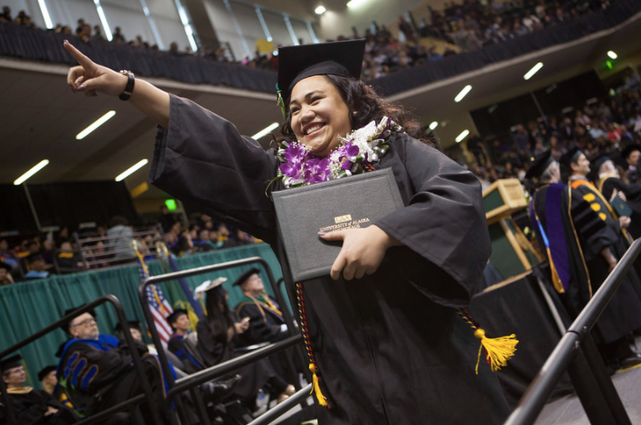 A woman graduate walking across the stage in regalia points up into the crowd
