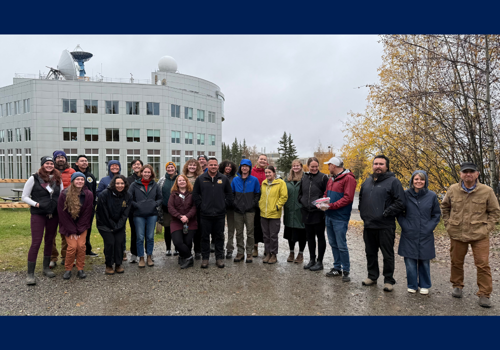 A large group of people stand outdoors on a cloudy day in front of the Akasofu Building at UAF, posing for a group photo with autumn trees in the background.