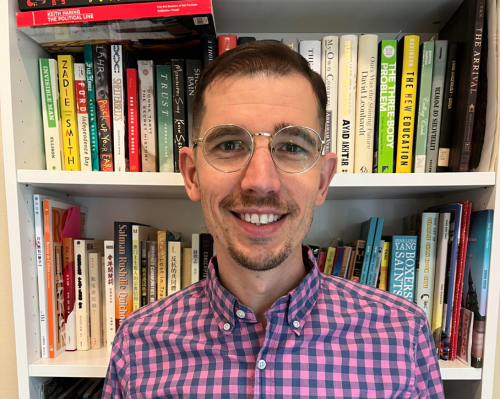 a man with glasses and plaid shirt smiles in front of a full bookshelf