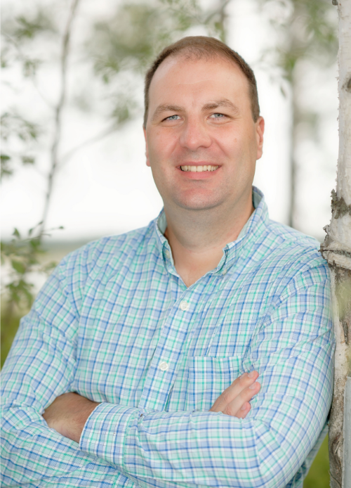 Headshot of Brandon Boylan, a man with short brown hair wearing a blue and green plaid shirt, smiling with arms crossed and leaning against a birch tree outdoors.