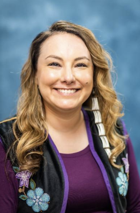 Woman with blonde hair and black vest smiles in front of a blue portrait background