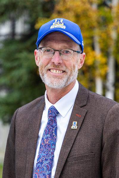 White wears a baseball cap and blue suit, stands in front of fall foliage