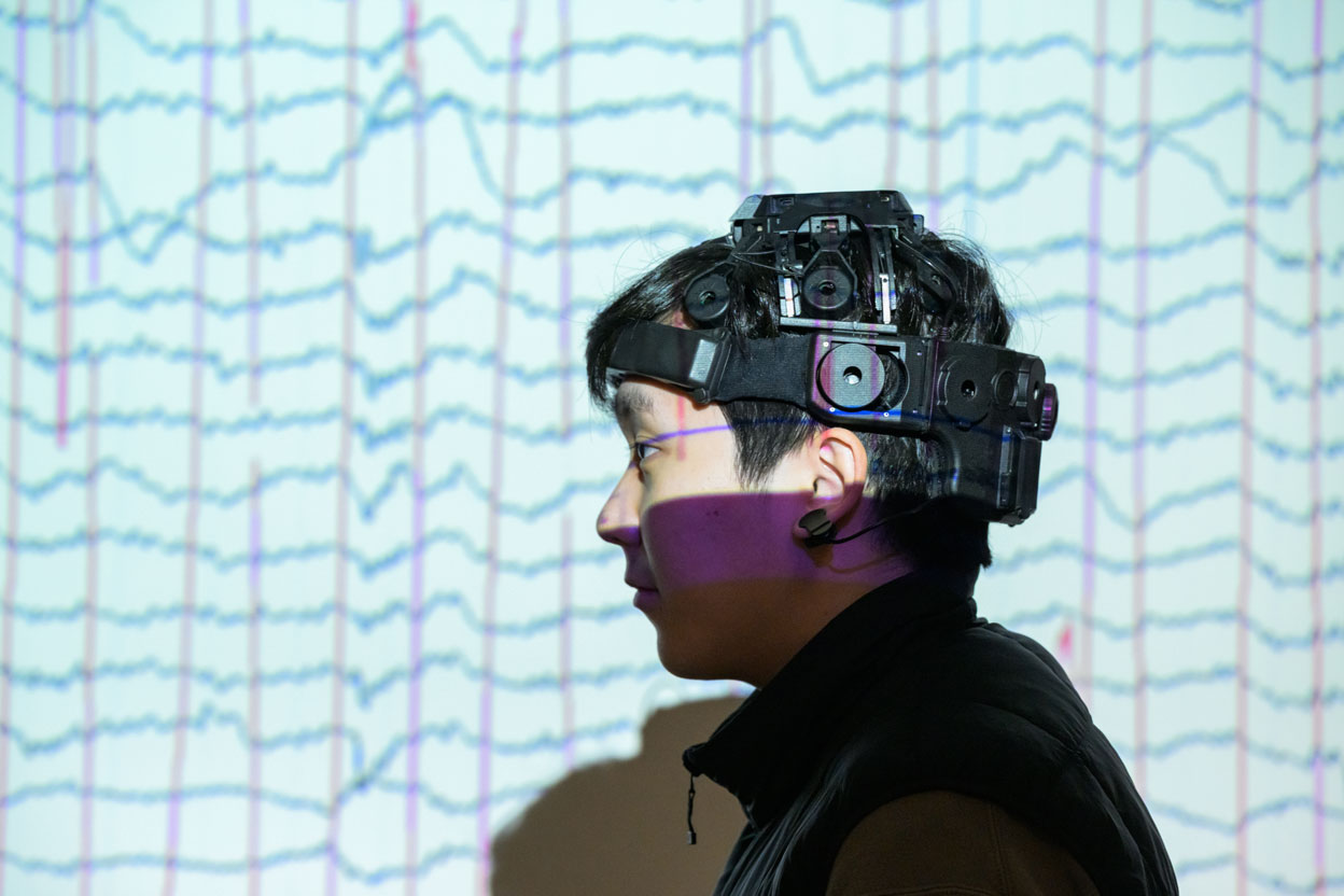 Side-profile view of a research participant wearing a specialized EEG headset with multiple sensors and cameras. Brainwave data graphs are projected across the background and partially onto the participant's face, illustrating neuroscience or brain-computer interface research.