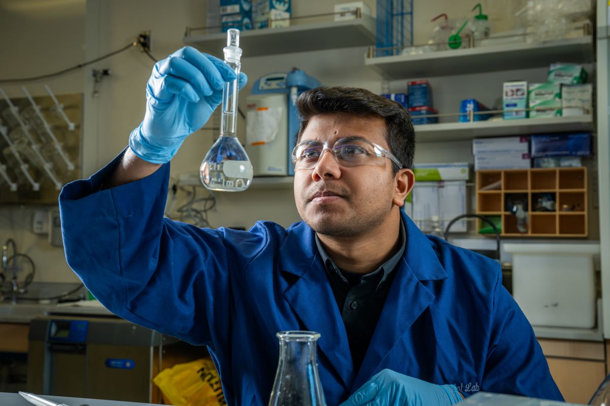 A man in a blue lab coat holds a glass beaker in front of his face