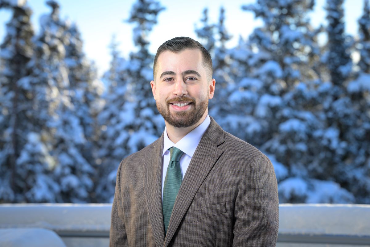A man in a grey suit and green tie in front of snow covered trees