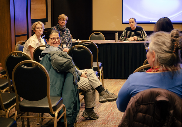 A small group of University of Alaska faculty and staff participate in a discussion during a workshop session. Several people sit in a semi-circle, listening and engaging in conversation in a conference room.