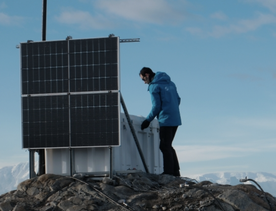 A man in a blue jacket stands on a rock mound next to four solar panels and a white box