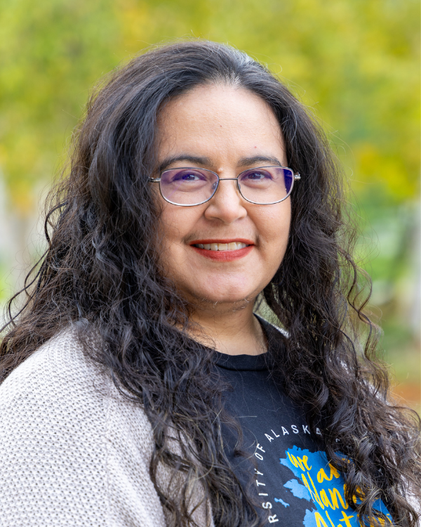 A woman with long curly hair, a tshirt and brown cardigan smiles in front of a green background.