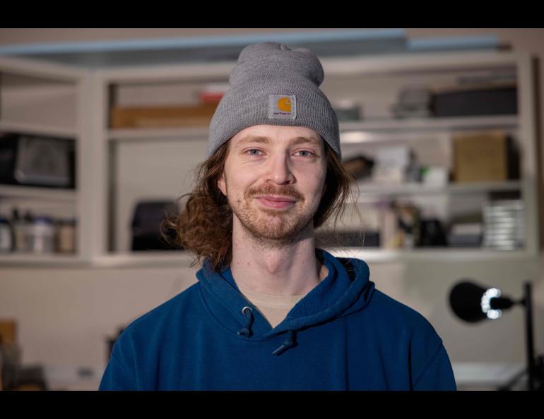 A young man with facial hair and grey beanie smiles at the camera from inside a lab
