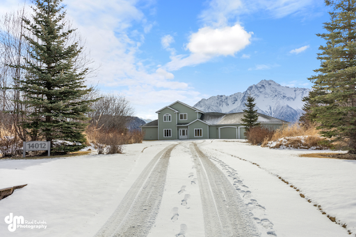 View of a large home at the end of a snowy driveway, with spruce trees and mountain range behind the house